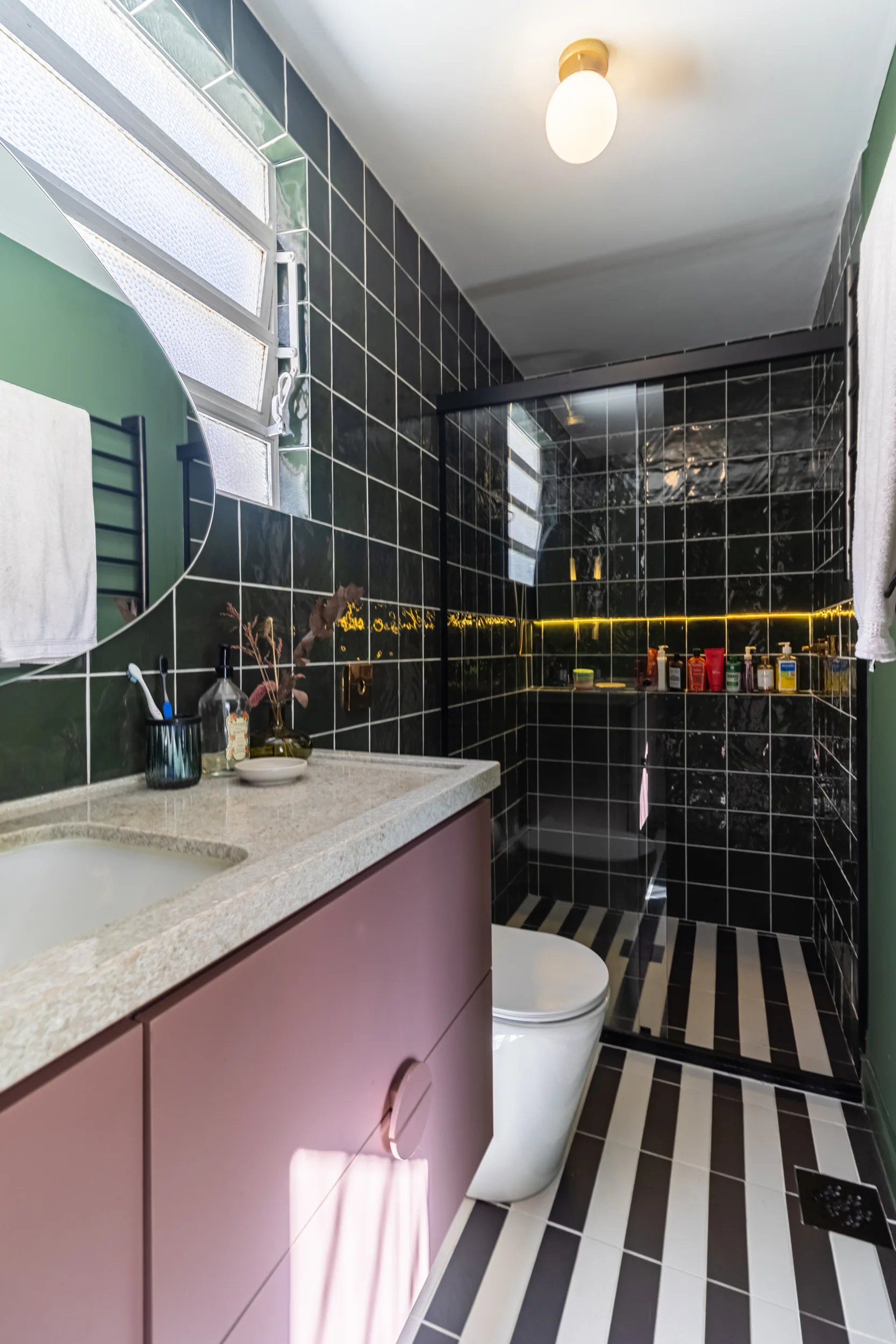 três vistas de um banheiro com azulejos pretos, marcenaria verde e rosa, espelho orgânico e piso listrado